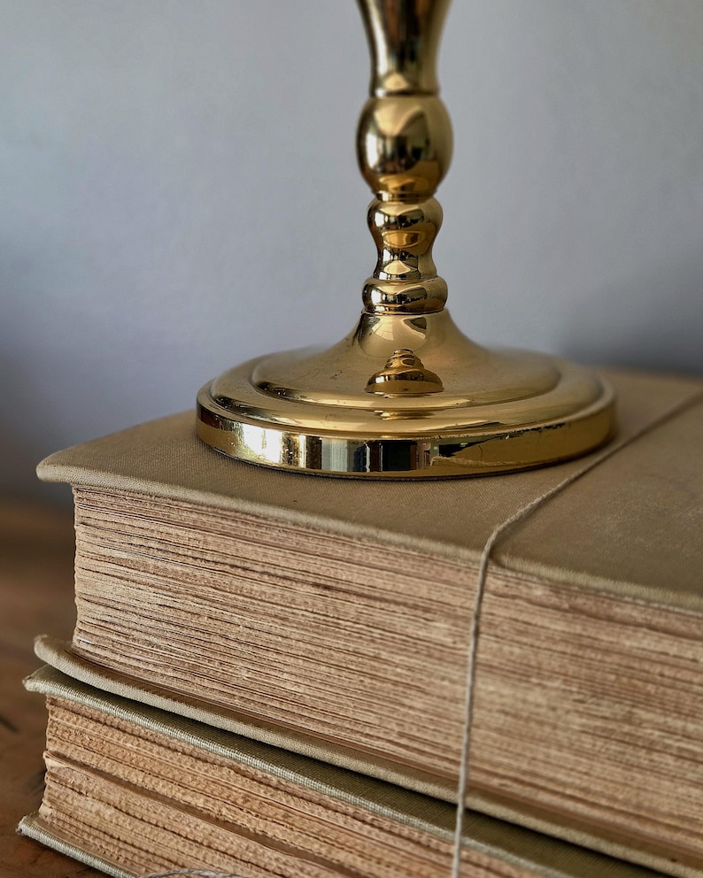 May include: A polished gold-toned metal candlestick base with a circular pedestal and a tiered, rounded stem, resting on top of two stacked, thick, beige-colored hardcover books with visible paper edges and a thin string wrapped around them.