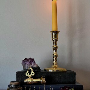 May include: A polished brass candlestick holding a lit yellow taper candle, positioned atop two stacked black books. Beside the candlestick sits a raw purple amethyst crystal, a brass key-shaped object, a small glass inkwell, and a dried dark rose.