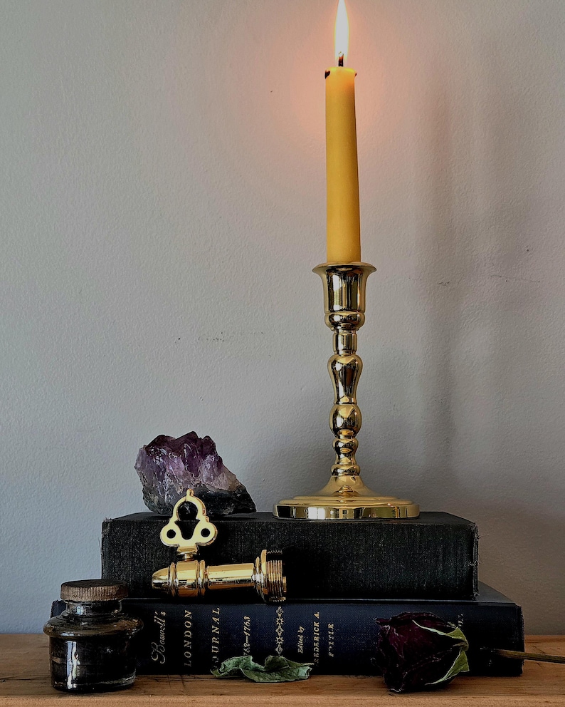 May include: A polished brass candlestick holding a lit yellow taper candle, positioned atop two stacked black books. Beside the candlestick sits a raw purple amethyst crystal, a brass key-shaped object, a small glass inkwell, and a dried dark rose.