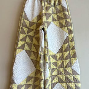 May include: Quilted pants with a geometric pattern. The pants feature a patchwork design with white, yellow, and brown triangles and squares. The waistband is elasticized. The pants are hanging on a wooden hanger against a neutral wall.