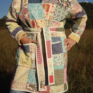 May include: A patchwork robe with a belt, featuring a variety of fabric patterns and colors. The robe has a quilted appearance with a mix of floral, geometric, and solid fabric squares. The belt is white with a patterned edge. The robe is worn over blue jeans.