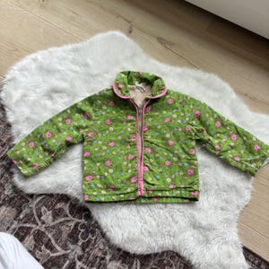Peut inclure: Une veste à manches longues verte avec un motif floral répétitif de fleurs roses et bleues. La veste présente une fermeture éclair frontale sur toute la longueur avec un liseré rose le long du col et de la fermeture, et une doublure en polaire claire.