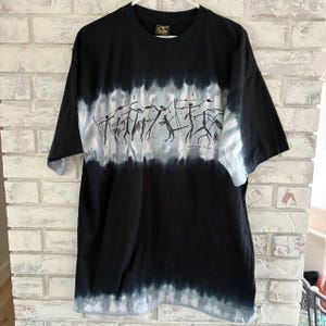 May include: A black short-sleeve t-shirt featuring a horizontal tie-dye band in shades of white and gray across the chest. The band displays a black graphic print of stick figures and a kangaroo. The shirt has a crew neckline and a black hem.