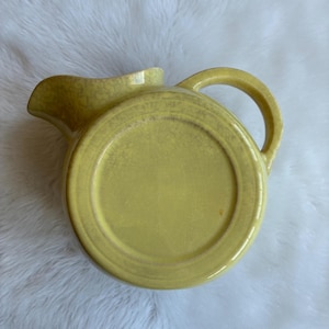 May include: A yellow ceramic pitcher with a handle and spout. The pitcher has a round base and a slightly textured surface. The handle is arched, and the spout is curved. The pitcher is likely vintage.