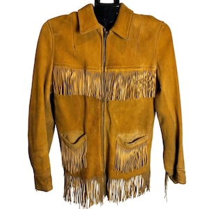 May include: A tan suede jacket with fringe detailing. The jacket features a zip-up front, two front pockets, and fringe along the chest, pockets, and hem. The jacket has a collared neckline and long sleeves.