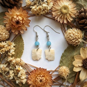 May include: A pair of dangle earrings with silver hooks, light blue beads, and off-white, rectangular charms. The earrings are displayed on a white surface, surrounded by dried flowers and pine needles.