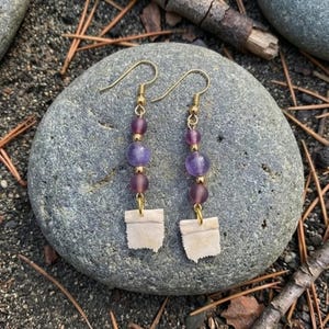 May include: A pair of dangle earrings with gold-colored hooks. Each earring features a combination of purple and mauve beads, with a small, rectangular, off-white charm at the bottom. The earrings are displayed on a gray stone.