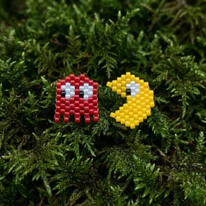 以下が含まれることがあります： ビーズで作られた2つのパックマンのフィギュア。1つは白と黒のディテールを持つ赤いゴースト、もう1つは白と黒の目を持つ黄色のパックマンです。フィギュアは緑の苔を背景に配置されています。