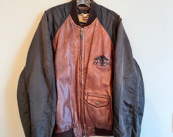 May include: A brown leather bomber jacket with black nylon sleeves. The jacket has a zipper closure and a patch on the left chest with the Harley Davidson logo and the text "1903 Harley Davidson 1983".
