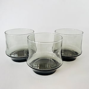 May include: Three gray glass tumblers with a black rim. The glasses are clear and have a slightly rounded shape.