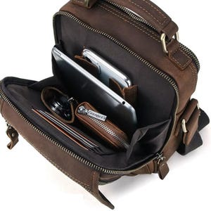 Può includere: Borsa a tracolla in pelle marrone con più scomparti, tra cui una sezione principale con cerniera e tasche esterne. La borsa è aperta, rivelando un tablet, un telefono, un portafoglio e altri oggetti. Ha un manico superiore e una tracolla regolabile.