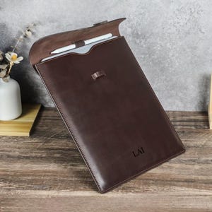 May include: A brown leather laptop sleeve with a flap closure. The sleeve has a stitched border and a small tab closure. A white stylus and a laptop are visible inside. The initials "LAI" are engraved on the front.