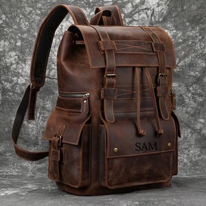 May include: A brown leather backpack with multiple pockets, straps, and buckles. The backpack has a drawstring closure and a flap with buckles. The name "SAM" is embossed on the front pocket. The bag has padded shoulder straps.