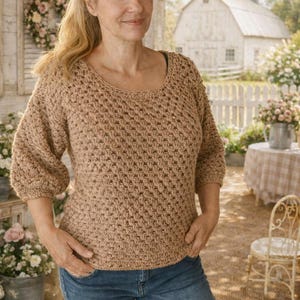 Può includere: Una donna indossa un maglione all'uncinetto color beige con scollo tondo e maniche a tre quarti, abbinato a jeans blu. Il maglione presenta un motivo a punto strutturato. Lo sfondo include una staccionata bianca, fiori e un fienile bianco.