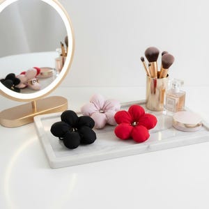 Peut inclure: Trois chouchous en forme de fleur, noirs, roses et rouges, disposés sur un plateau en marbre. Un miroir de maquillage doré et des pinceaux de maquillage sont en arrière-plan. Un flacon de parfum et un poudrier sont également sur le plateau.