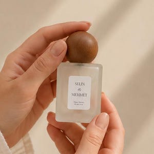 May include: A square, frosted glass perfume bottle with a round, dark wooden cap. The front label features the text SELIN & MEHMET with smaller text below. The bottle is held upright, showcasing its minimalist design and smooth, matte texture.