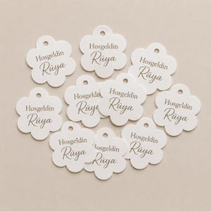 May include: A collection of ten white, flower-shaped tags, each featuring a small hole at the top. The text 'Hosgeldin Rüya' is printed in a dark gray, elegant serif and script font on the center of each individual tag.