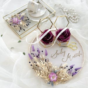 May include: A wedding ring holder with a floral arrangement and two hexagon-shaped ring boxes. The ring boxes contain burgundy roses and are tied with white ribbons. A tray with a teacup and saucer is also present.