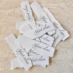 May include: A collection of white, rectangular paper strips with deckled edges. Each strip features the black printed text Afiyet olsun in a cursive script. The paper has a textured, fibrous appearance, and the strips are arranged in a scattered pile.