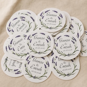 May include: A stack of circular white paper stickers featuring a printed lavender wreath design. Each sticker displays the names Gamze & Emrah in a black script font, with the date 08.08.2026 printed in black below the names.