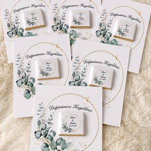 May include: Six white square chocolate favors attached to matching white cards. Each card features a gold circular frame, green eucalyptus leaves, and the text Düğünümüze Hoşgeldiniz. The chocolates are printed with Merve & Atakan 12.10.2027.
