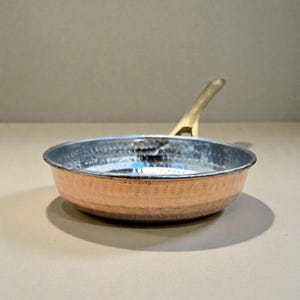 May include: A small copper frying pan with a hammered texture and a brass handle. The interior is a silver color. The pan is designed for cooking and is likely used for preparing individual servings or delicate dishes.