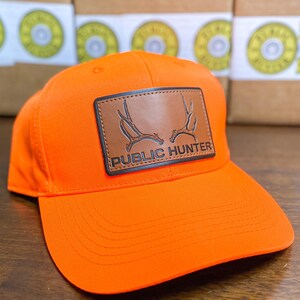 May include: Bright orange baseball cap with a brown leather patch. The patch features a deer antler design and the words "PUBLIC HUNTER" in white. The cap has a curved brim and a structured fit.
