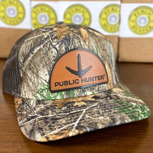 May include: Camouflage baseball cap with a brown leather patch featuring a black turkey footprint and the words "PUBLIC HUNTER". The cap has a mesh back and a curved brim. The hat is designed for outdoor activities.
