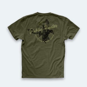 May include: Olive green t-shirt with a graphic of a duck in flight and the words "Public Hunter" in a stylized font. The text "Feet Down, Beat Down" is below the duck. The shirt has a crew neck and short sleeves.