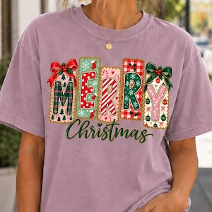 May include: A mauve short-sleeved t-shirt featuring a graphic print of the word MERRY in colorful, patterned block letters decorated with bows, holly, and candy canes, with the word Christmas written in green cursive script underneath.
