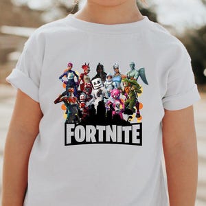 May include: White t-shirt featuring a colorful graphic of various Fortnite characters. The word "FORTNITE" is printed in bold, black letters below the characters. The shirt is being worn by a child.