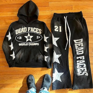 Könnte beinhalten: Ein schwarzer Kapuzenpullover und eine passende Jogginghose mit weißen, abgenutzten Sternenmotiven und Schriftzügen. Der Hoodie zeigt "DEAD FACES WORLD CHAMPS" mit "20" und "21". Die Hose ist mit "21" und "DEAD FACES" sowie weißen Kordeln versehen.