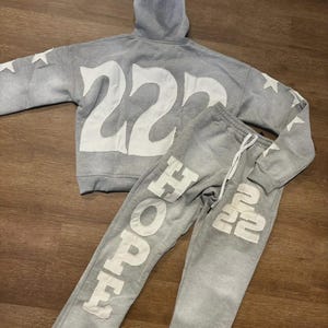 Könnte beinhalten: Ein graues Hoodie- und Jogginghosen-Set. Der Hoodie hat die Nummer "222" in Weiß, und die Jogginghose sagt "HOPE 222" in Weiß. Weiße Sterne befinden sich auf den Ärmeln des Hoodies.