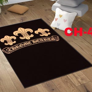 Può includere: Un tappeto rettangolare nero con tre simboli giglio color oro disposti orizzontalmente sopra la scritta CHROME HEARTS in un carattere gotico stilizzato. Il tappeto presenta uno sfondo scuro a tinta unita con il disegno centrato nella parte alta.