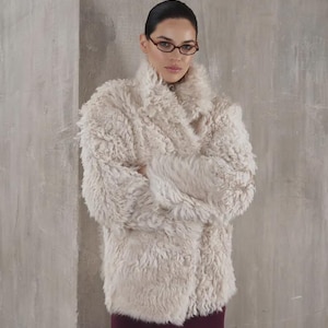 Könnte beinhalten: Ein cremefarbener Kunstfellmantel mit breitem Kragen und Wickelverschluss. Der Mantel wird mit einem bordeauxfarbenen Rock kombiniert. Das Model trägt eine Brille. Der Hintergrund ist eine neutrale graue Wand.