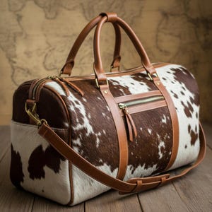 May include: A cowhide duffel bag in brown and white, with tan leather detailing. It has a top handle, a shoulder strap, and several zipped compartments. The metal hardware is gold-coloured. This travel bag has a classic, rustic design.