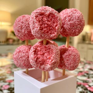 May include: A collection of pink cake pops, each coated in a layer of pink sprinkles. The cake pops are arranged in a white square container, with wooden sticks inserted into each one. The treats are a vibrant pink color.