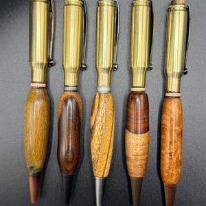 May include: Five handcrafted pens with brass bullet casings and wooden barrels. The barrels feature various wood grains and colors, including light and dark browns, and a striped pattern. Each pen has a metal clip and a different colored tip.