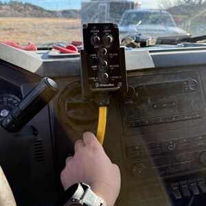May include: A black control panel with the text "SYSTEM", "LIGHTS", "LOAD", "UNLOAD", "GRIP", and "RELEASE". The panel is mounted on a vehicle dashboard. A yellow hose is connected to the panel. A person's hand is holding the hose.