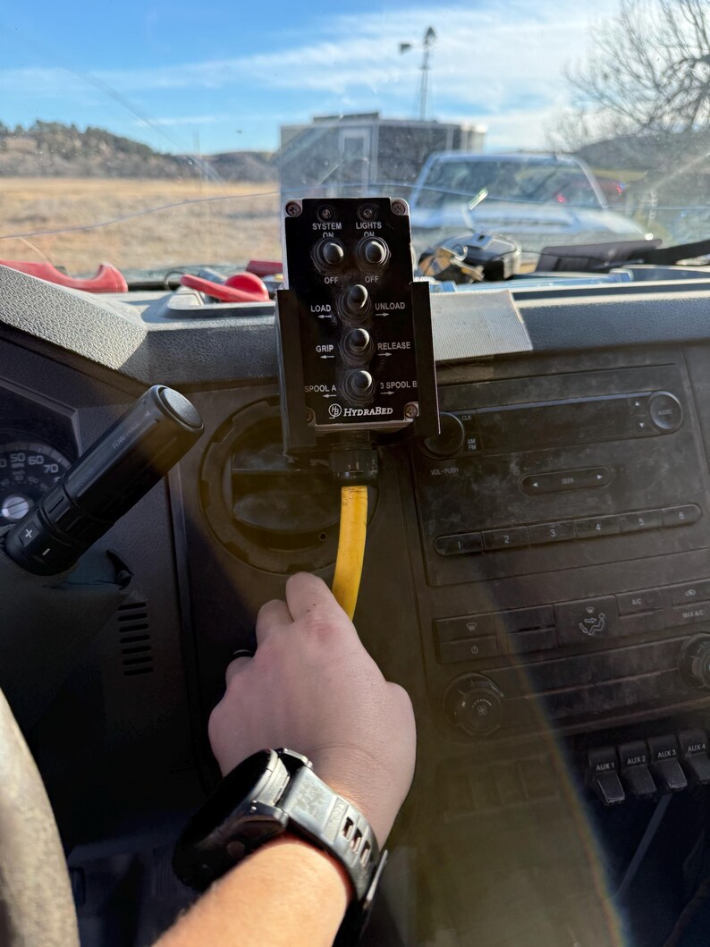 May include: A black control panel with the text "SYSTEM", "LIGHTS", "LOAD", "UNLOAD", "GRIP", and "RELEASE". The panel is mounted on a vehicle dashboard. A yellow hose is connected to the panel. A person's hand is holding the hose.