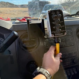 May include: A control panel with multiple switches and the text "SYSTEM", "LIGHTS", "LOAD", "UNLOAD", "GRIP", "RELEASE", "3 SPOOL A", and "SPOOL B". The panel is mounted on a vehicle dashboard. A hand is holding a yellow hose.