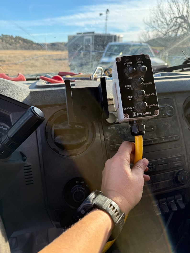 May include: A control panel with multiple switches and the text "SYSTEM", "LIGHTS", "LOAD", "UNLOAD", "GRIP", "RELEASE", "3 SPOOL A", and "SPOOL B". The panel is mounted on a vehicle dashboard. A hand is holding a yellow hose.