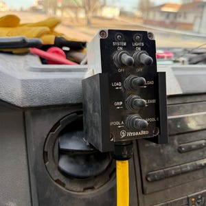 May include: A black control panel with multiple switches and the text "SYSTEM ON", "LIGHTS ON", "LOAD", "GRIP", "LEASE", "SPOOL A", and "SPOOL B". A yellow hose is connected to the bottom of the panel. The brand name "HYDRABED" is also visible.