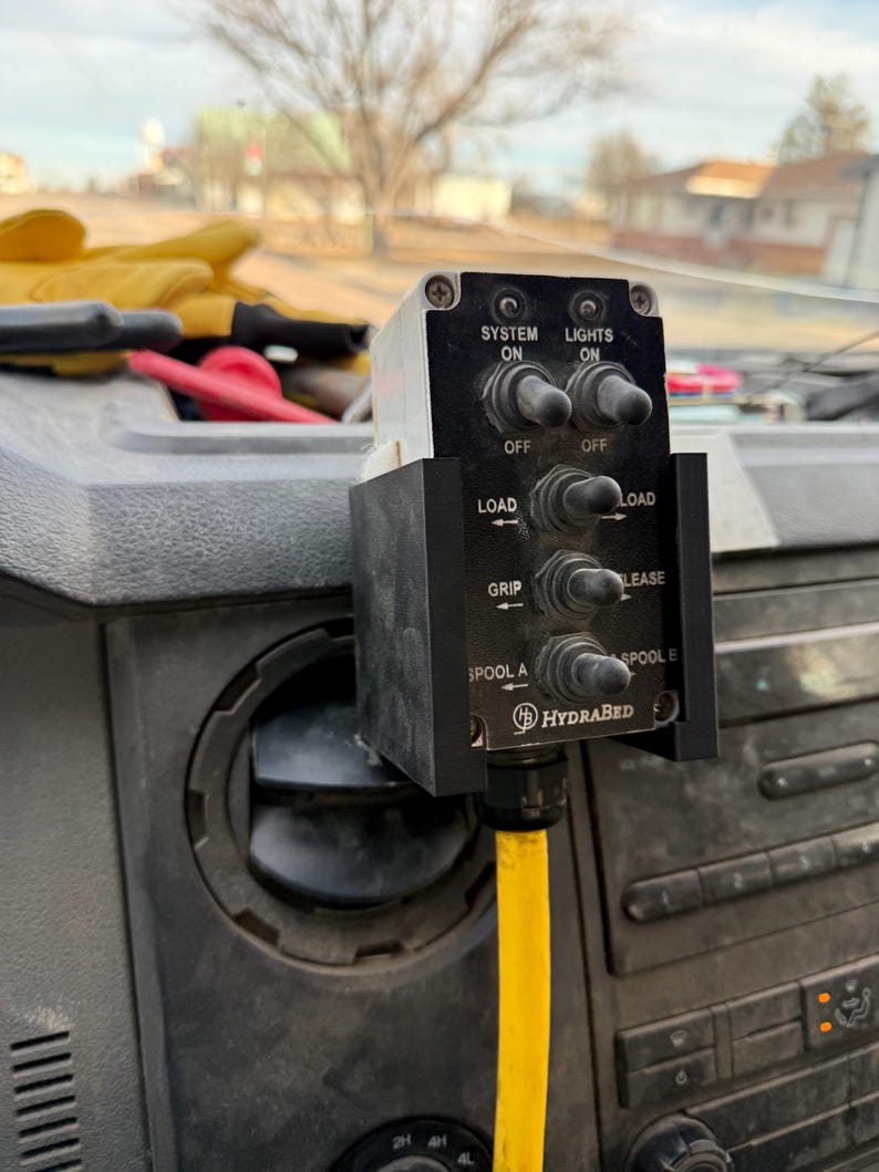 May include: A black control panel with multiple switches and the text "SYSTEM ON", "LIGHTS ON", "LOAD", "GRIP", "LEASE", "SPOOL A", and "SPOOL B". A yellow hose is connected to the bottom of the panel. The brand name "HYDRABED" is also visible.