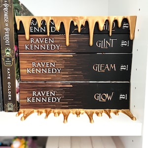 May include: A gold-colored bookend with a dripping design holds a stack of books. The books have black covers with the title "Raven Kennedy" and other titles. The bookend is on a white shelf with other books visible.