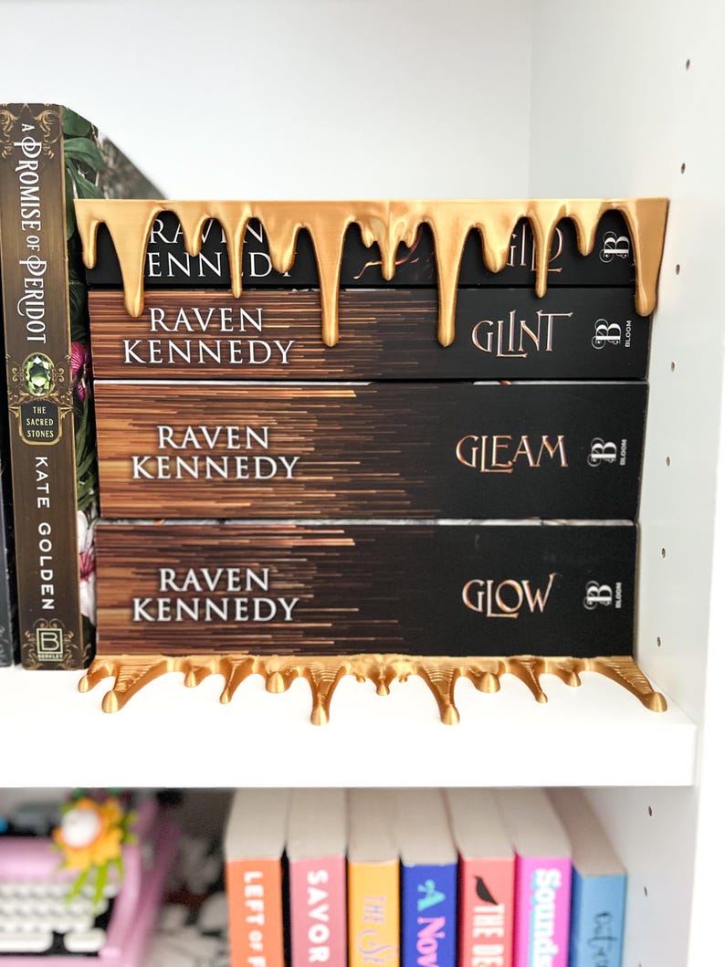 May include: A gold-colored bookend with a dripping design holds a stack of books. The books have black covers with the title "Raven Kennedy" and other titles. The bookend is on a white shelf with other books visible.