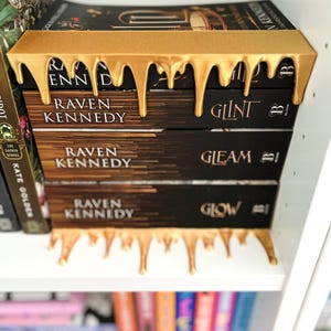 May include: A stack of three books by Raven Kennedy, with gold-colored decorative bookends. The bookends have a dripping paint design. The book titles are "Glint" and "Gleam" and "Glow". The books are on a white shelf.