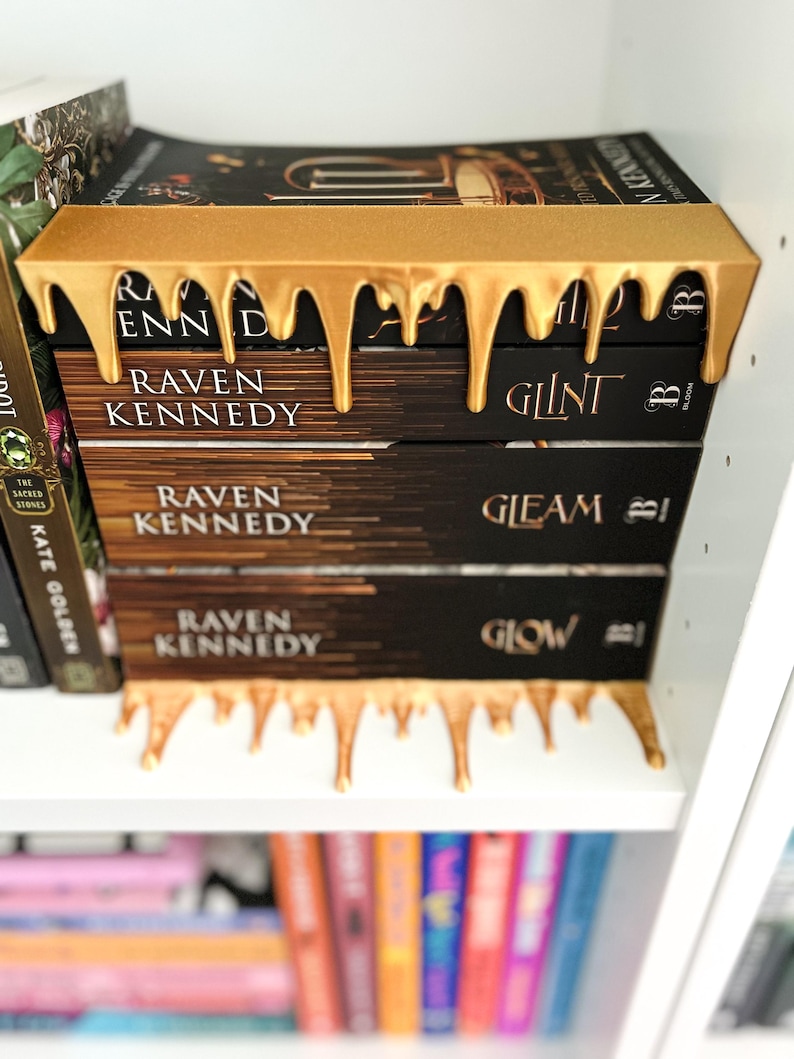 May include: A stack of three books by Raven Kennedy, with gold-colored decorative bookends. The bookends have a dripping paint design. The book titles are "Glint" and "Gleam" and "Glow". The books are on a white shelf.
