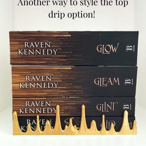 May include: A stack of three books with black covers and gold lettering, titled "Glow", "Gleam", and "Glint" by Raven Kennedy. A gold-colored, stylized drip design holds the books in place. The text "Another way to style the top drip option!" is at the top.
