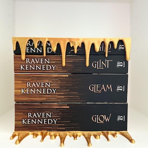 May include: A stack of three books with black covers and gold lettering, titled "Glint", "Gleam", and "Glow" by Raven Kennedy. The books are held by a gold-colored decorative shelf with a dripping paint design.